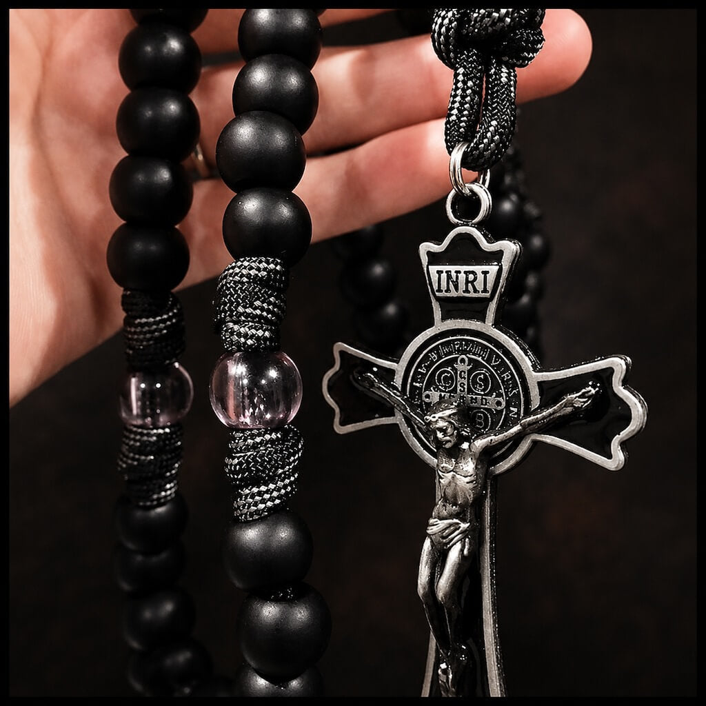 Adult hand holding the Black Monk rosary to show the size of its matte beads, knotted cord, and St. Benedict crucifix.